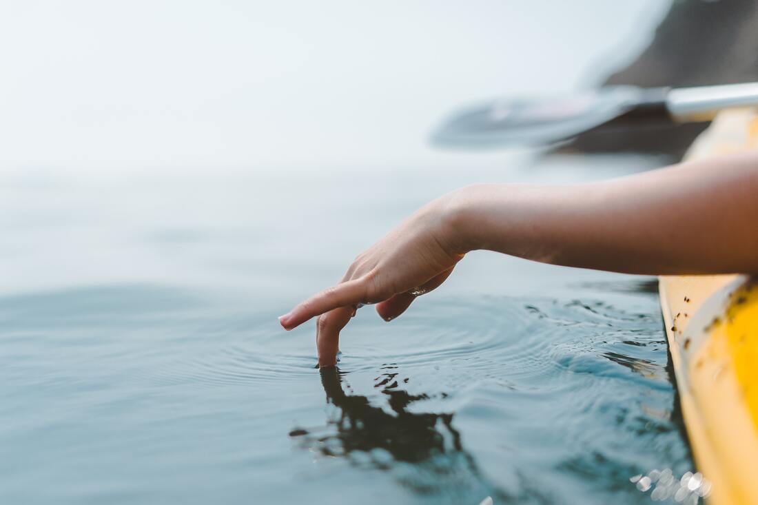 Fotografia da superfície da água, talvez o mar. Do lado direito, um braço estica-se de uma canoa amarela, e a mão toca a água muito suavemente, com apenas um par de dedos a tocá-la. Ao fundo, vê-se a pagaia e atrás uma falésia, ambas desfocadas.