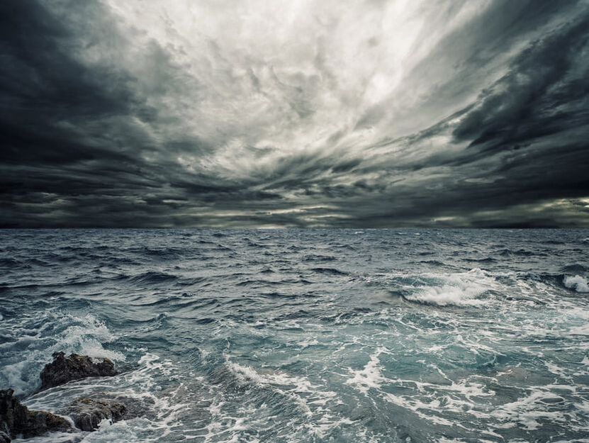 Fotografia de uma tempestade iminente. O mar agitado em tons de cinzento, com ondas e algumas rochas em baixo à esquerda ocupa a metade de baixo da fotografia. Na metade de cima está o céu carregado com nuvens negras. O céu abre um pouco ao centro, onde as nuvens são mais claras e passa alguma luz.