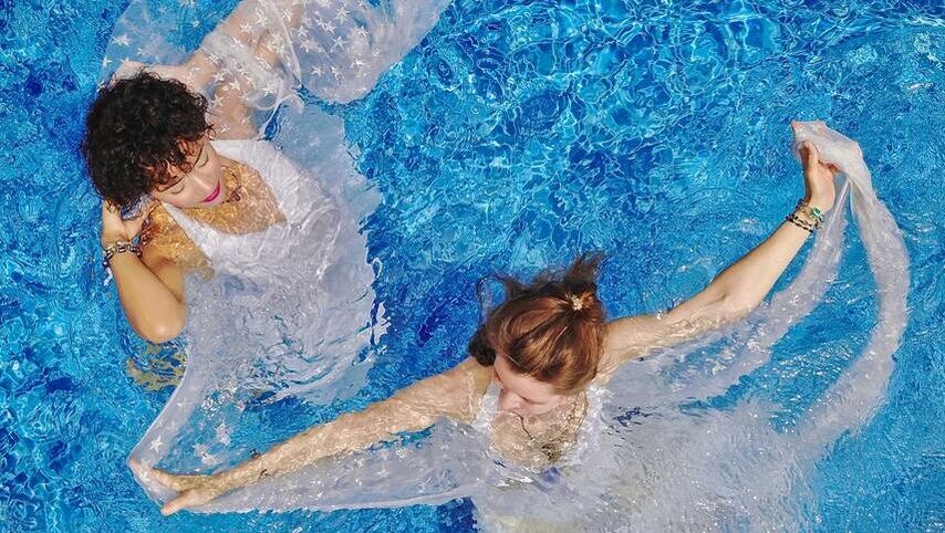Vista de cima de duas mulheres com vestidos brancos que parecem estar a dançar dentro de uma piscina com fundo azul.