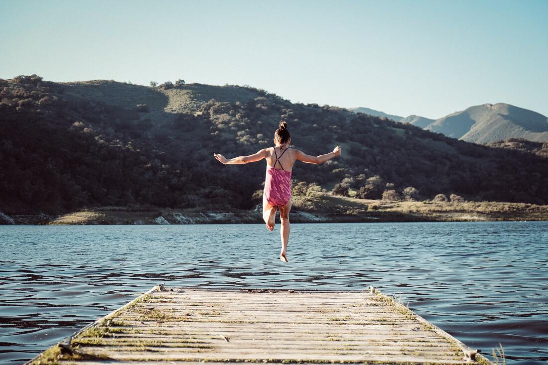 Uma criança salta para um lago a partir de um pequeno cais de madeira. A menina está de costas, veste um fato de banho cor-de-rosa e tem os braços abertos. Ao fundo vê-se a outra margem do lago com colinas cobertas de vegetação. O céu está limpo e pelas sombras percebe-se que o Sol brilha.