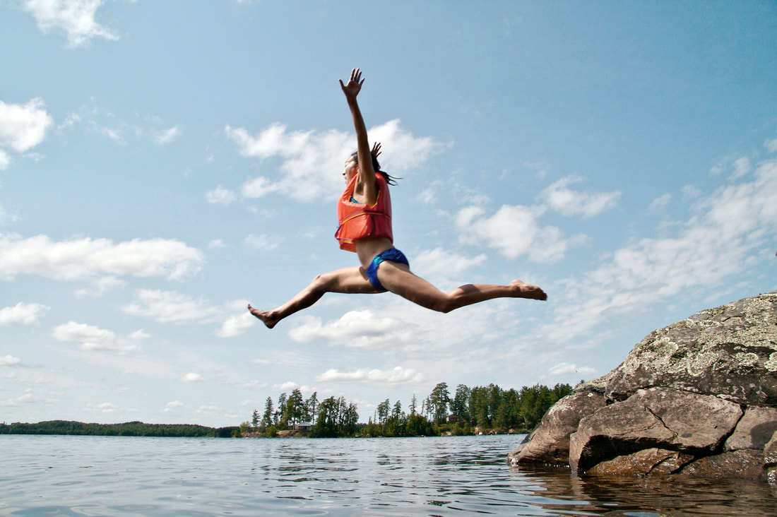 Um homem a meio de um salto com o céu em fundo. À direita está uma rocha e em baixo o lago para onde ele salta. Ele tem um colete salva-vidas laranja, fato-de-banho azul e tem as pernas e os braços abertos.