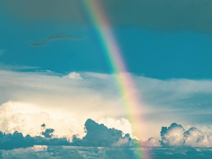 Imagem do céu com nuvens na parte de baixo e o céu azul na metade de cima. Um arco-íris atravessa o céu de alto a baixo.