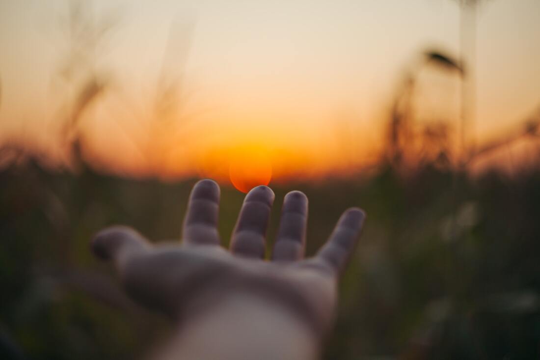 Uma mão está esticada para a frente com a palma virada para cima. O sol laranja põe-se ao fundo, aparentemente tocando a ponta dos dedos. O resto do fundo está desfocado e mostra alguma erva alta verde escura e o céu alaranjado.