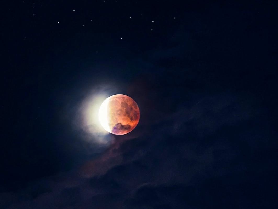 Fotografia de um eclipse lunar. Uma sombra avermelhada cobre a maior parte da lua cheia, excepto uma ponta do lado esquerdo, que está muito iluminada. O fundo é um céu azul escuro profundo com algumas nuvens debaixo da lua e algumas estrelas por cima dela.